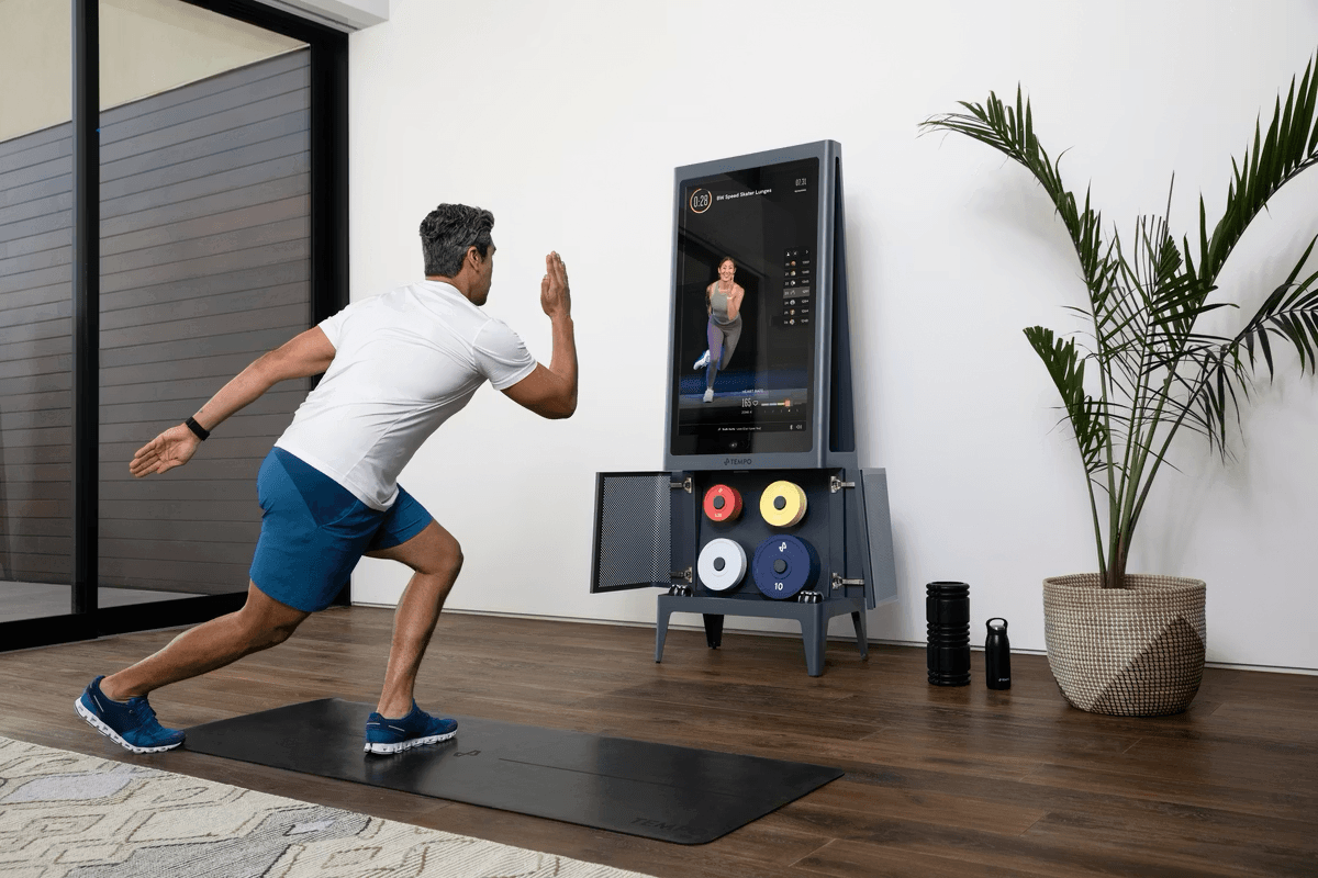 Man doing lunges on a mat in front of a Tempo smart home gym showing a live class